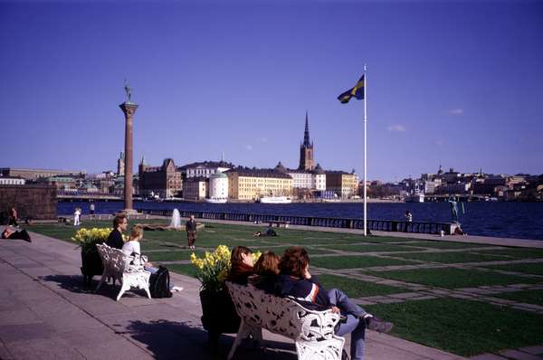 photo of Sweden, Stockholm, couple sitting on white bench in the garden of the Stockholm City Hall (stadshuset) enjoying the view of Gamla Stan (Old Town) and the Swedish flag