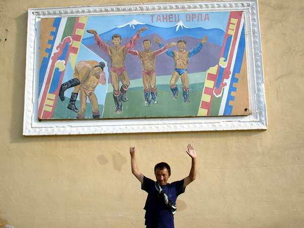 photo of Tuva, Tuvan man with one hand in front of a painting of a traditional wrestling competition. As the wrestlers are introduced to the crowd before the match, they do the dance of the eagles, where they wave their hands like an eagle wings; the winner will also get to do the dance of the eagle around the flag pole after the match