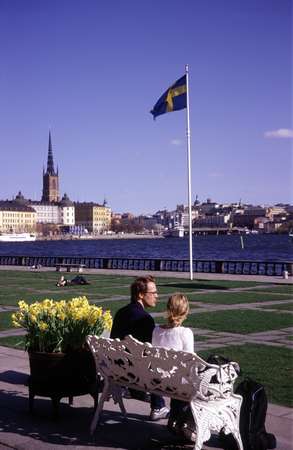photo of Sweden, Stockholm, couple sitting on white bench in the garden of the Stockholm City Hall (stadshuset) enjoying the view of Gamla Stan (Old Town) and the Swedish flag
